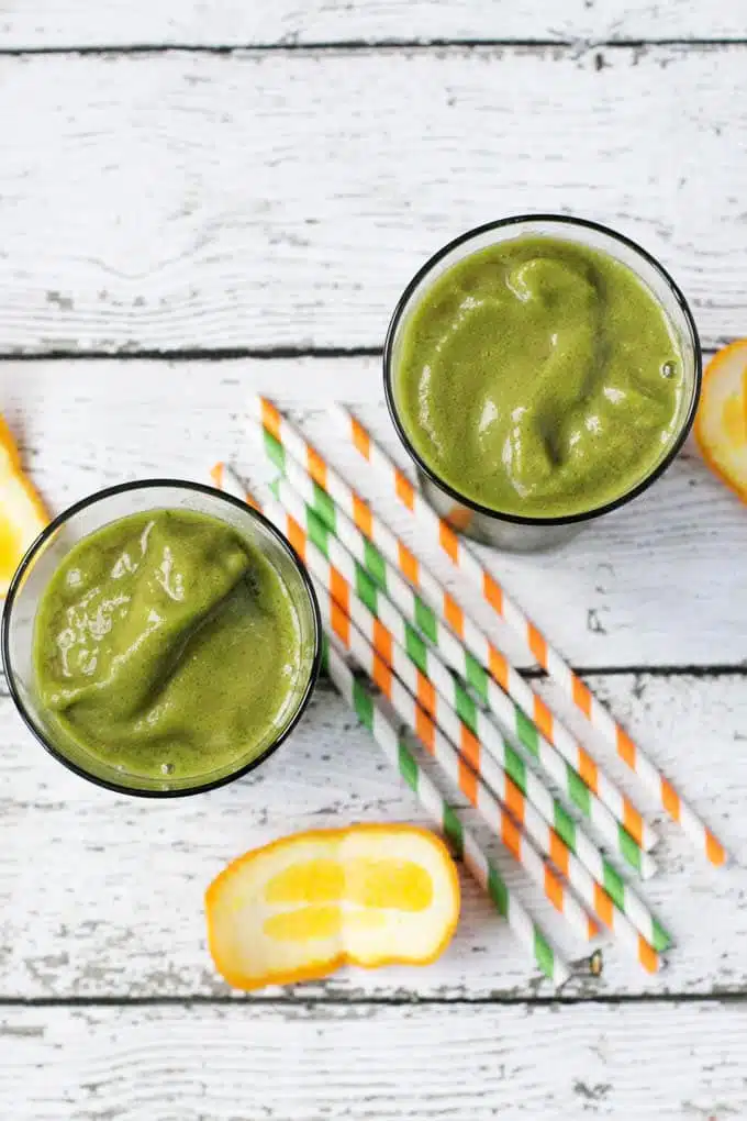 top down view of two glasses of green smoothie surrounded by straws and orange peels