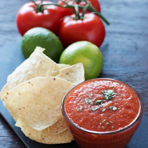 cup of salsa on dark background with tortilla chips, fresh tomatoes and green tomatillas