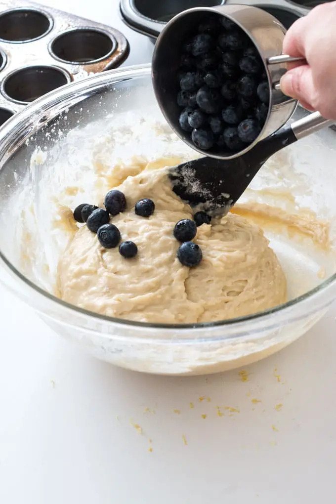 A cup of blueberries being poured into bowl of batter.