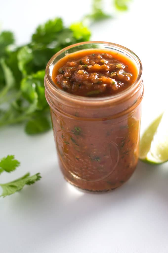 A jar of salsa on a white background surrounded by fresh cilantro and a slice of lime.