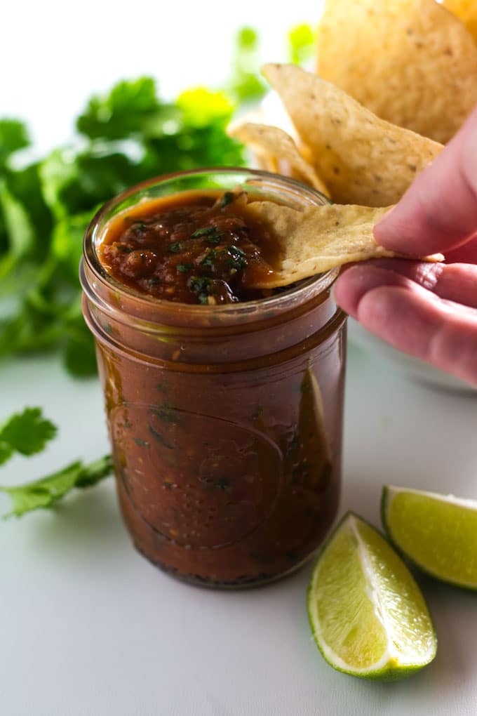 a hand dipping a tortilla chip in a jar of salsa on a table surrounded by lime slices, fresh cilantro and a bowl of tortilla chips.