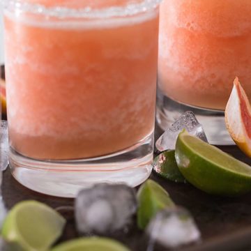 Two glasses of frozen paloma cocktail on a metal sheet pan surrounded by ice and lime wedges