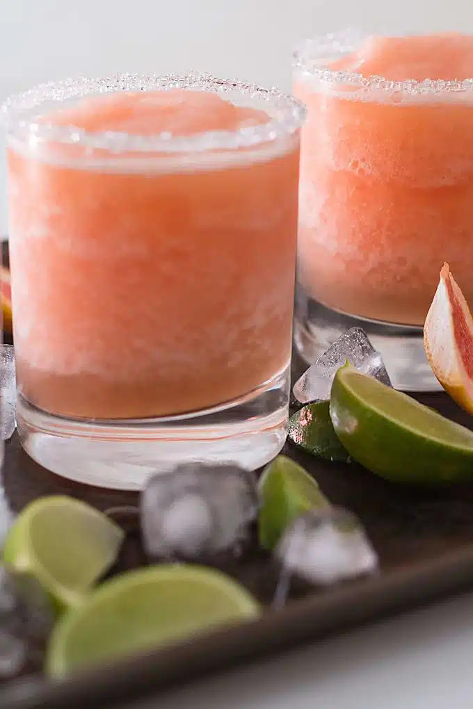 Two glasses of frozen paloma cocktail on a metal sheet pan surrounded by ice and lime wedges